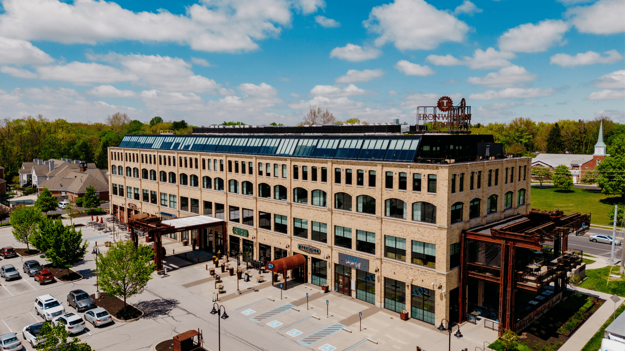 Aerial view of Ironworks Indy, a light brick four-story building on a clear day with puffy clouds and vibrant green trees. Ironworks Hotel Indy has been named #9 Top Hotel in the United States.