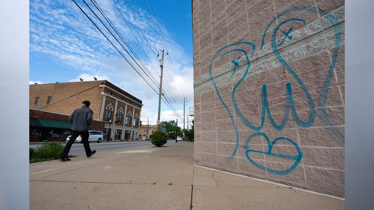 A pedestrian walks past graffiti on the side of CSL Plasma in Irvington on May 10, 2024. The face-like depiction has been left by an anonymous artist in multiple locations around the east-side neighborhood. (Photo by Jenna Watson/Mirror Indy)