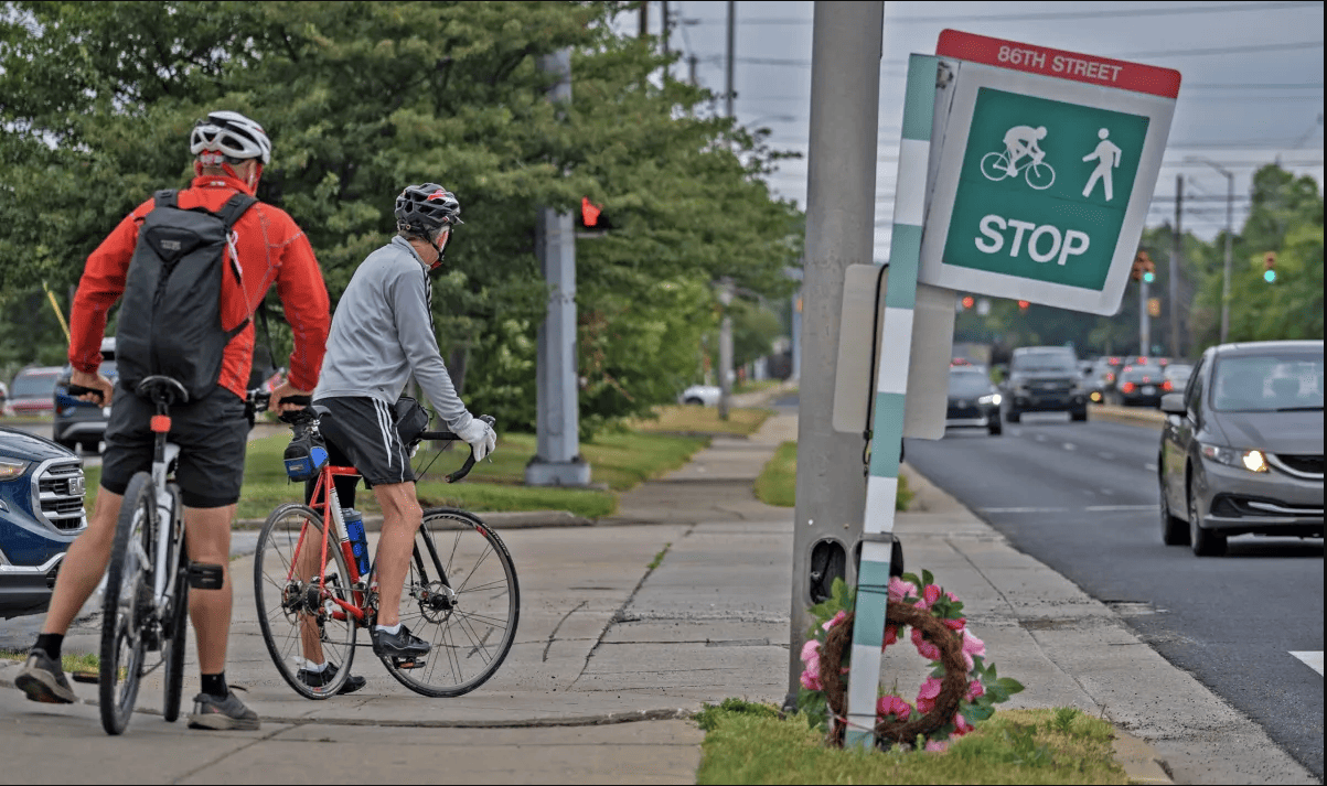 Cyclists get ready to cross 86th Street to continue their path on the Monon Trail on June 12, 2023, on the north side of Indianapolis. A wreath memorializing Frank Radaker is seen at right near the crosswalk.