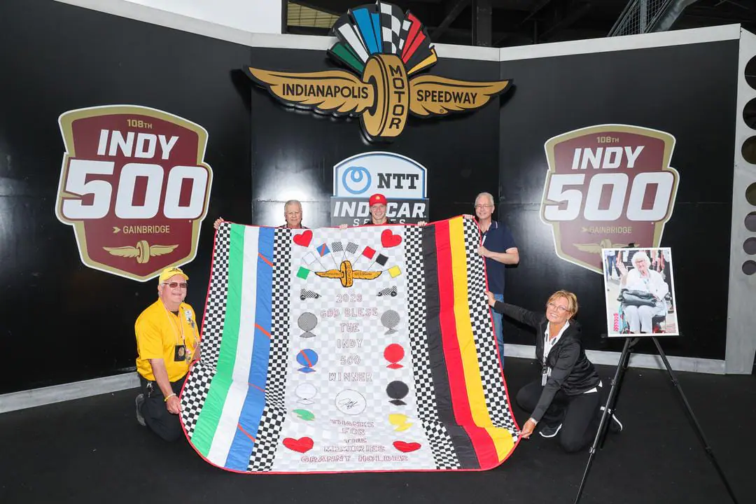 2023 Indianapolis 500 winner Josef Newgarden (center) helps hold up the quilt made for him by the late Jeanetta Holder with help from Holder's family and others. Holder, known by drivers and fans as the 